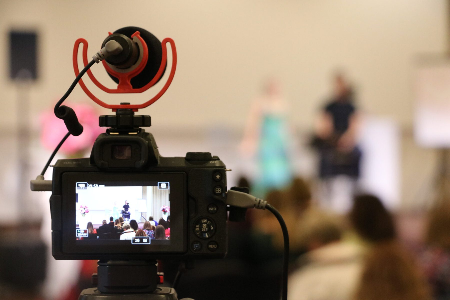Professional mirrorless camera with a microphone filming a live ceremony, the seated audience visible on the camera screeny in a hall