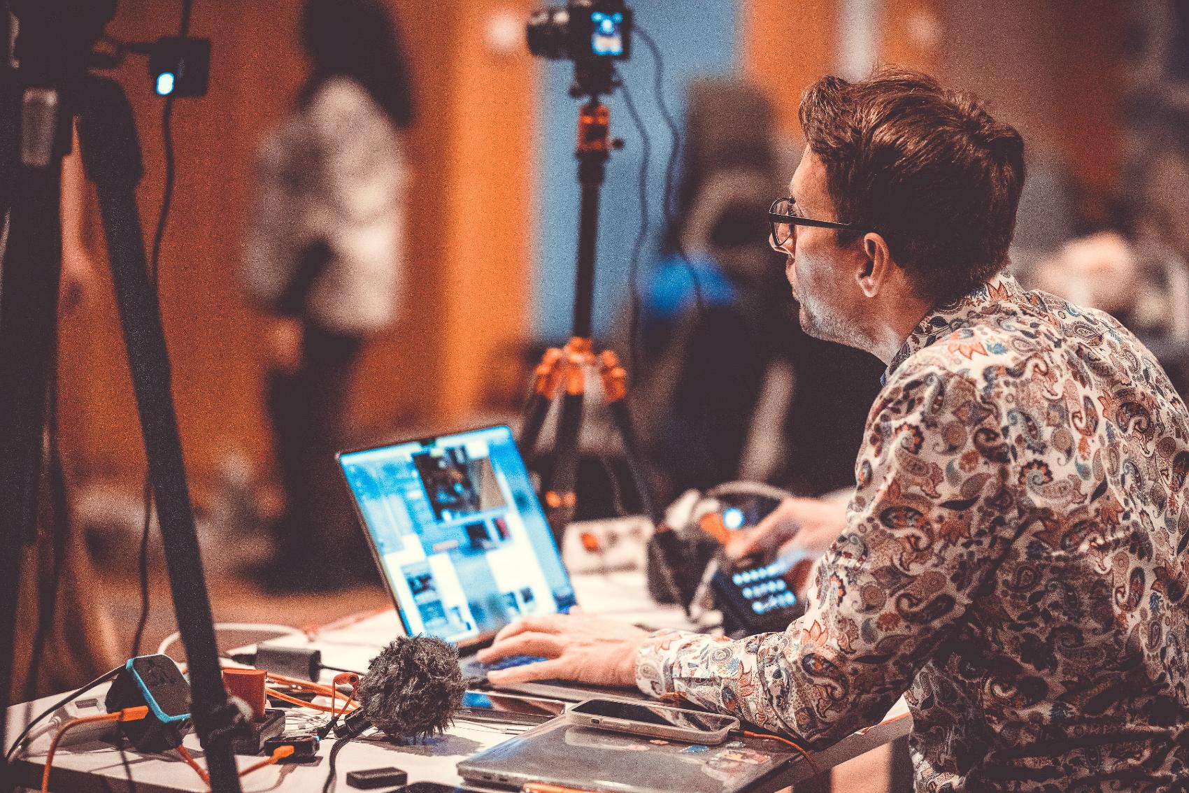 Livestream operator at a laptop running multi-camera streaming software with cameras and microphones at a live eventuple on stage in front of a seated audience and floral arrangements control on a laptop with a stream deck